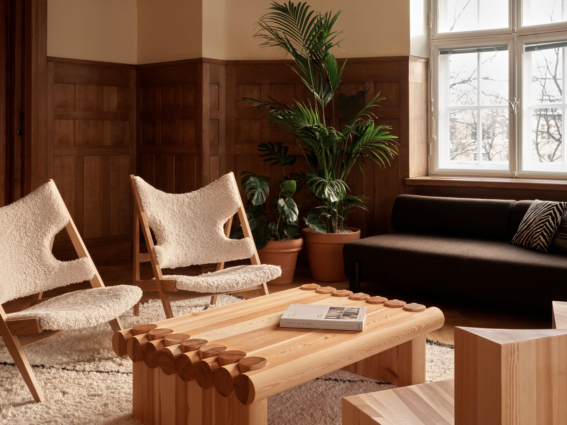 wooden panels, solid wood crafted furniture, large coffee table, dark grey-brown sofa, couple of lounge chairs, large plants, warm atmosphere